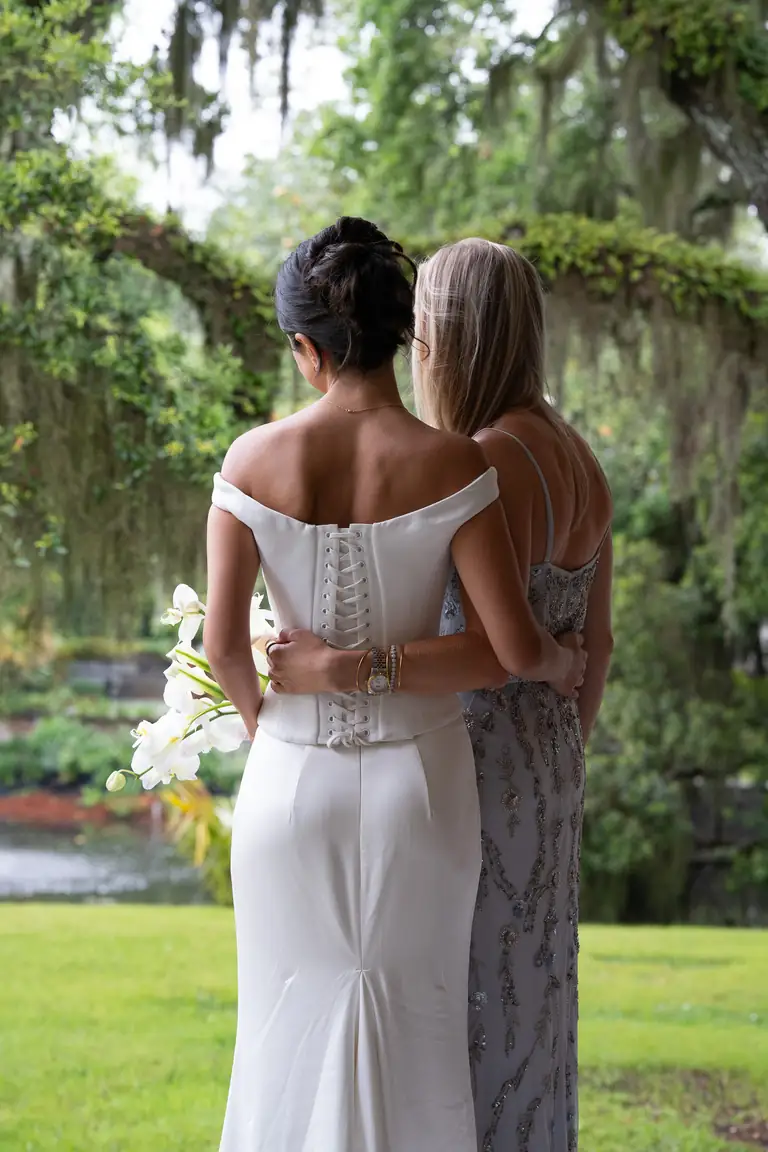 A bride and a guest embracing on the lawn beneath moss-draped oaks