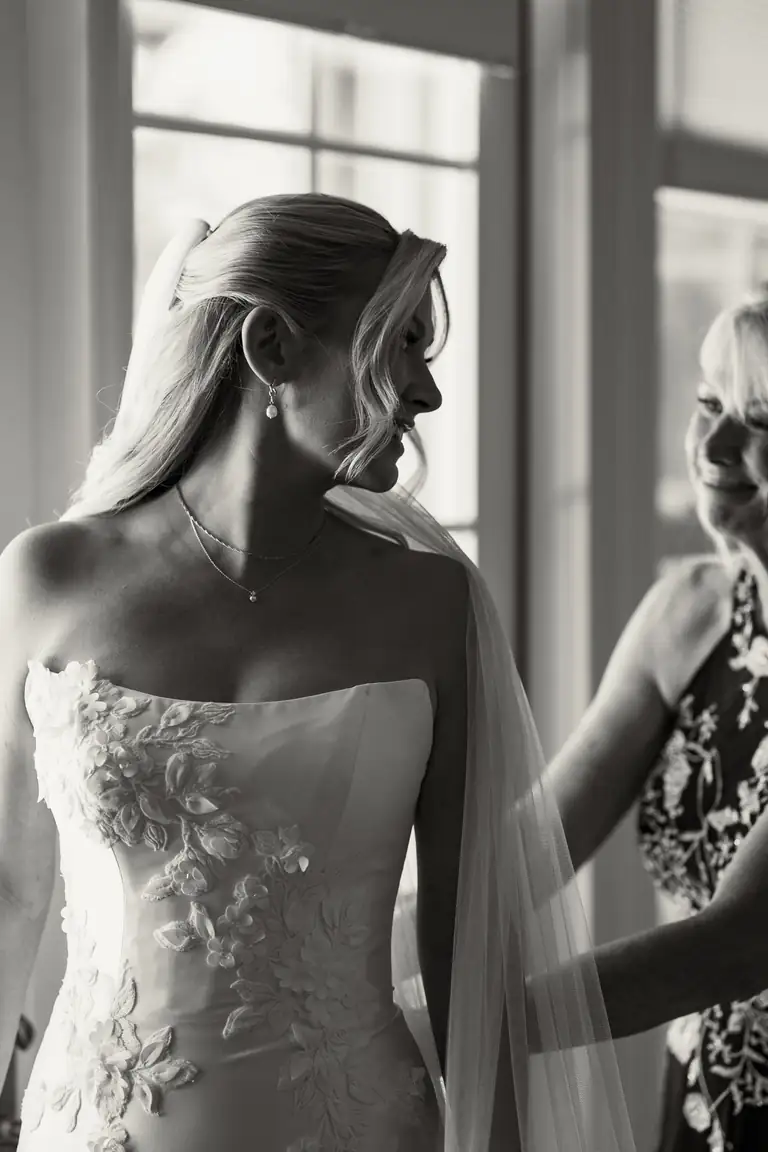 A bride in a beaded gown being helped into it by the window, black and white