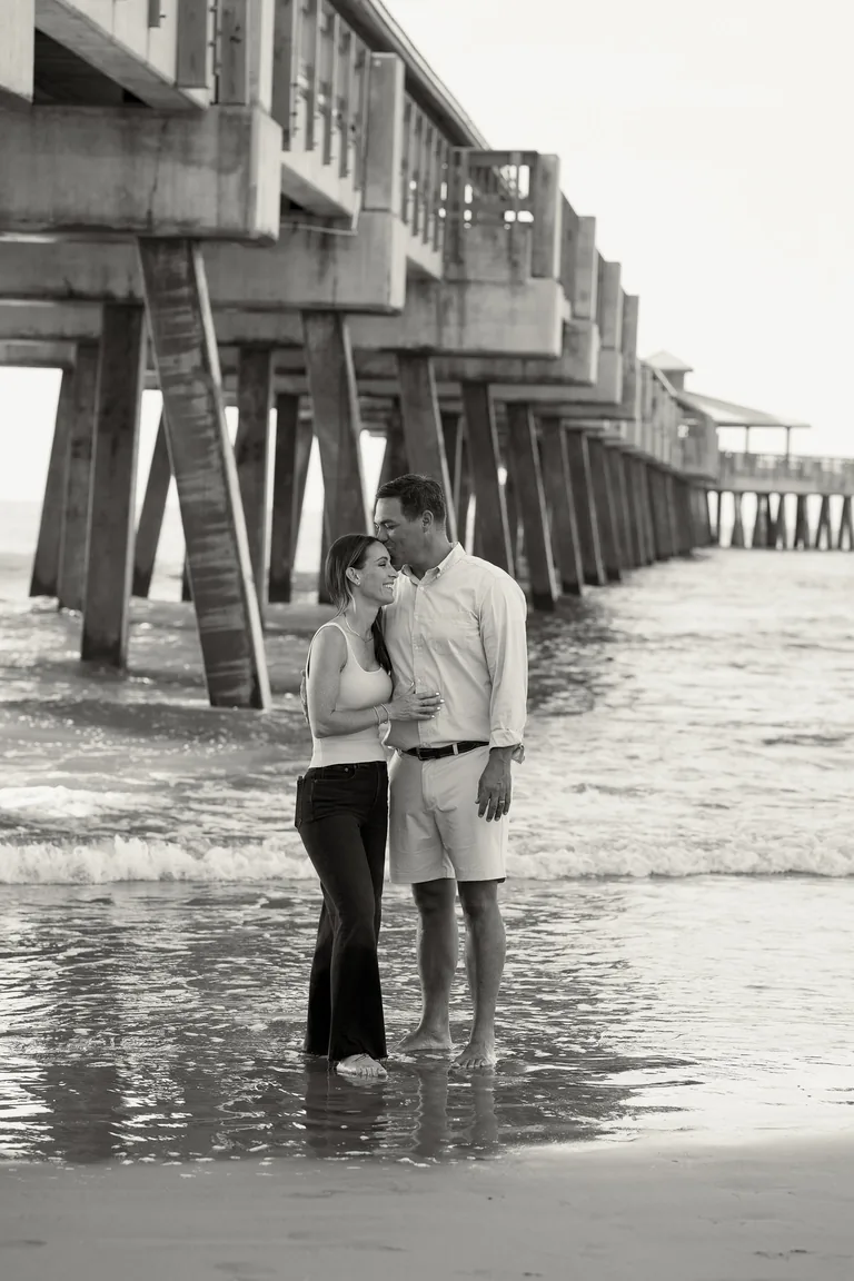 A couple standing together on wet sand beneath a pier, black and white